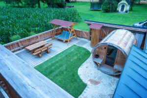 Einladender Außenbereich eines Holzhaus‑Chalets mit gepflegter Garten‑ und Pflasterfläche, Holzzaun, Holz‑Terrassendielen und einer rustikalen Fasssauna vor natürlicher Kulisse von ERLER  Haus.