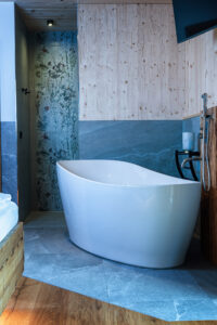 Ein Schlafzimmer mit freistehender Badewanne, Walk‑in‑Dusche und warmen Holz‑ sowie Fliesenelementen im Sanitärbereich von ERLER Haus.
