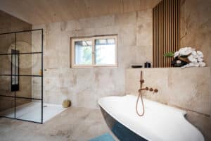 Ein modernes Badezimmer mit Walk‑in‑Dusche mit Glastrennwand, freistehender Badewanne, großformatigen Boden und Wandfliesen und einem Holzsprossen‑Wandelement von ERLER Haus.