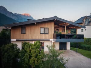 Ein Haus mit Satteldach, schwarzem Glasgeländer am überdachten Balkon, schwarzen Holz‑Alu‑Fenstern und Fassadenelementen aus Holz von ERLER  Haus.
