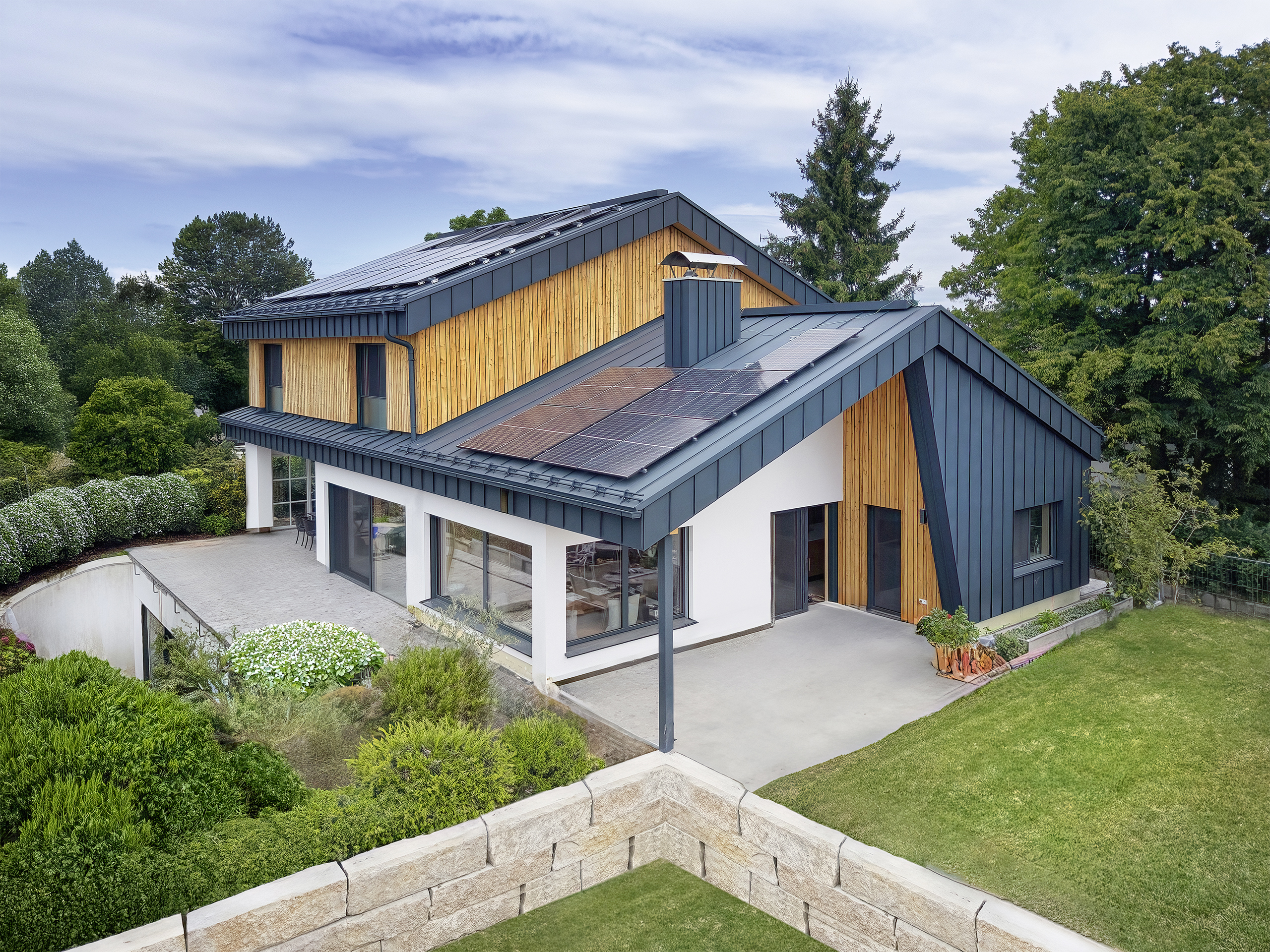 Ein mehrstöckiges Haus in Seitenansicht mit markanter Steinmauer, Holz‑Alu‑Fenstern, Fassadenelementen aus Holz und Blech und dachmontierten Solarpaneelen von ERLER Haus.
