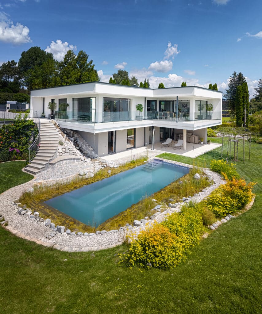 Außenansicht einer modernen Villa mit Flachdach, umlaufendem Balkon mit Glasgeländer, Aussenaufgang und einer Terrasse mit einem verspieltem Garten mit Pflasterung und einem in die Natur eingebetteten Pool von ERLER Haus.