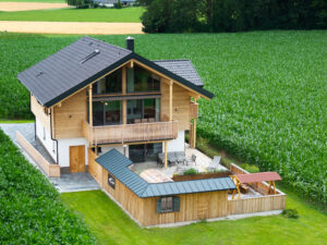 Ein Holzhaus‑Chaletbau aus der Vogelperspektive mit großem Holzbalkon, umlaufendem Zaun, gepflastertem Terrassenbereich und harmonischer Verbindung von Natur und Architektur von ERLER Haus.