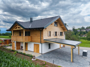 Seitenansicht eines Holzhauses mit Balkon, Carport, Wärmepumpe, Holz‑Alu‑Fenstern, rustikalen Fensterläden und gepflastertem Aussenbereich von ERLER Haus.