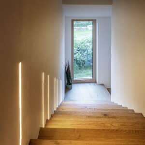 Ein markanter Treppenbereich mit Holztreppe, Podest, Glaselement und einer modernen wandintegrierten Lichtinstallation von ERLER  Haus.