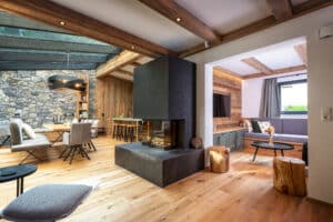 Ein offener Wohnraum mit offenen Nebenräumen, Decke mit sichtbaren Holzbalken und integrierten Spots, Wandelementen aus Holz und Stein sowie einem gemauerten Sichtkamin mit Sitzfläche von ERLER Haus.