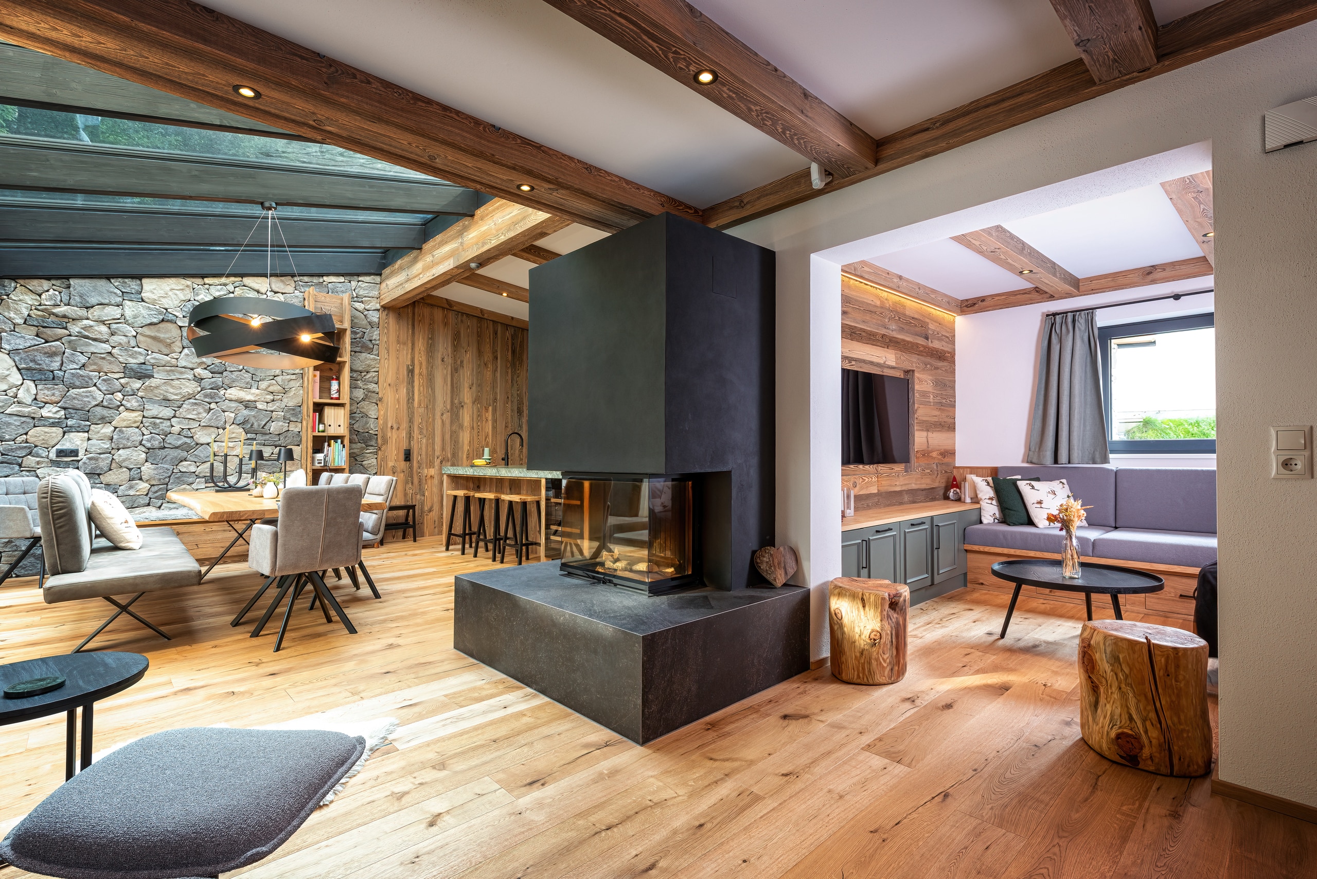 Ein offener Wohnraum mit offenen Nebenräumen, Decke mit sichtbaren Holzbalken und integrierten Spots, Wandelementen aus Holz und Stein sowie einem gemauerten Sichtkamin mit Sitzfläche von ERLER Haus.