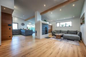 Ein offener Wohnraum mit Küche und durch einen gemauerten Kamin abgetrennten Wohnbereich, sichtbarem Deckenbalken, Deckenspots und harmonischer, gemütlicher Raumwirkung von ERLER Haus.