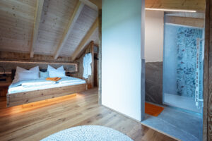 Ein großes Schlafzimmer mit Dachschräge, Holzbalken, Wandvertäfelung, und einem abgeteilten Badezimmer mit Walk‑in‑Dusche und indirekter Beleuchtung von ERLER Haus.