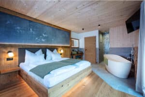 Ein großzügiges Schlafzimmer mit Holzdecke, Wandvertäfelung und grauen Steinfliesen im Waschbereich mit freistehender Badewanne, wandmontierter Flatscreen, Walk‑in‑Dusche und Holzwaschtisch mit aufgesetztem Steinwaschbecken von ERLER Haus.
