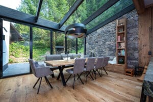 Ein offener Wohn- und Essbereich mit Wintergarten-Anschluss, Steinfassade, Terrassentüren und einem Glasdach mit dunklem Holzbalken von ERLER Haus.