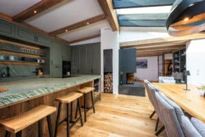 Ein offener Wohn‑ und Essbereich mit Wintergarten‑Anschluss, offener Wohnküche mit Theke in Grüntönen und einem gemauerten Kamin mit Holzlager von ERLER  Haus.