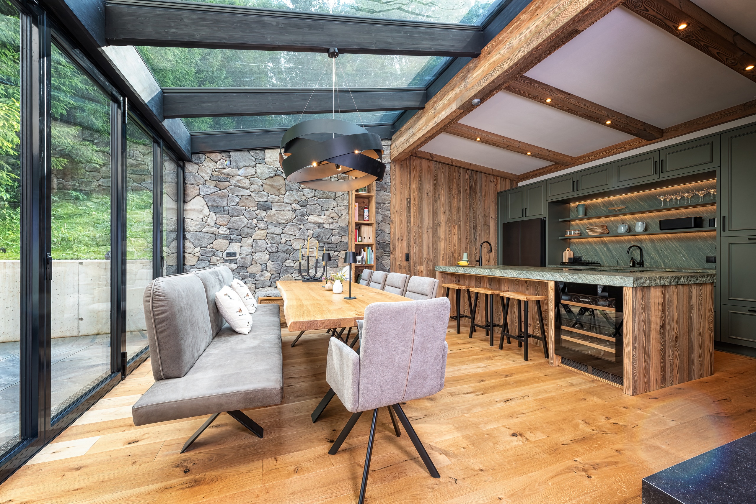 Ein offener Wohn‑ und Essbereich mit Wintergarten‑Anschluss, Steinfassade, offener Wohnküche mit Kochinsel, gemütlicher gepolsterter Sitzbank und Stühlen im Essbereich und Deckenbalken mit integrierten Spots von ERLER  Haus.