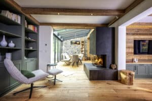 Ein offener Wohnraum mit offenen Nebenräumen, Holzbalken‑Decke mit Spots, gemauertem Kamin, integriertem Bücherregal und angrenzendem Wintergarten von ERLER  Haus.