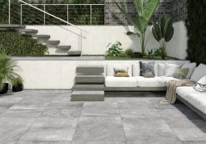 Fliesendorf zeigt eine Terrasse mit einer kleinen Begrünung im Eck, darunter eine weiße Lounge mit mehreren Polstern und die grauen Fliesen am Boden und auf den Treppen.