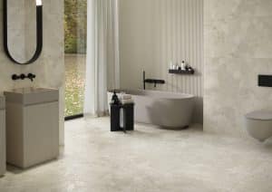 Fliesendorf zeigt elegantes Badezimmer mit großformatigen Bodenfliesen aus beigen Naturstein, freistehende cremefarbene Badewanne, Waschbecken und Toilette mit schwarzen Armaturen und Accessoires..