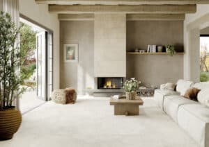 Wohnzimmer mit großformatigen hellen Steinfliesen an Wand und Boden aus der Portland-Rock-Reihe von Fliesendorf, kombiniert mit warmen Holzelementen, sichtbaren Deckenbalken und natürlichen Accessoires für einen gemütlichen, rustikalen und mediterranen Look.