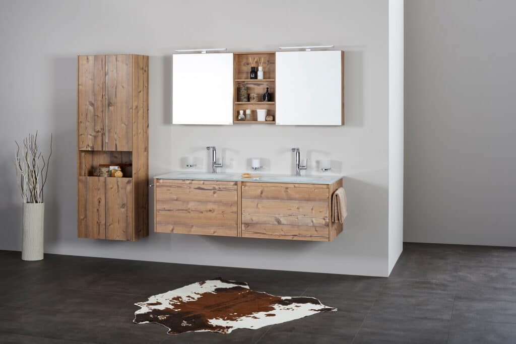 Frauenthal Handel zeigt ein Badezimmer mit Doppelwaschbecken, Unterschrank aus rustikalem Holz mit zwei großen Schubladen, Hochschrank und einem Spiegelschrank mit zwei Türen, offenen Ablagefächern und Beleuchtung.