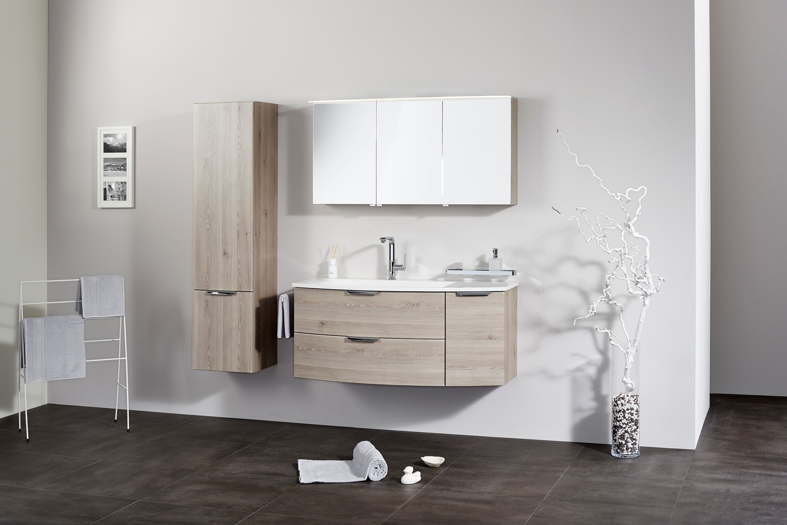 Frauenthal Handel zeigt ein Badezimmer mit einem Waschbecken, Unterschrank aus hellem Holz mit drei Schubladen, schmalen Hochschrank und einem Spiegelschrank mit drei Türen und Beleuchtung.