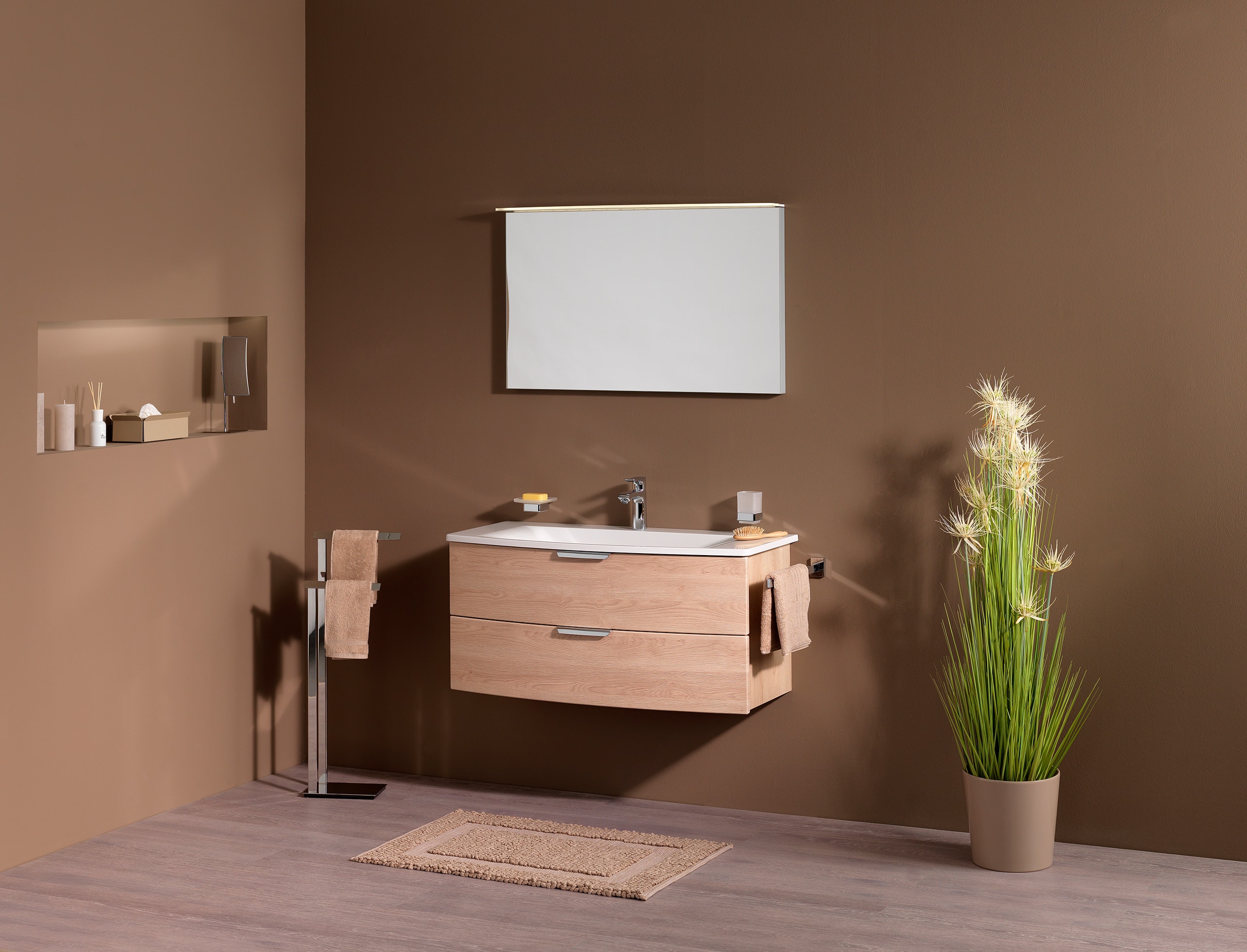 Frauenthal Handel zeigt ein Badezimmer mit einem Waschbecken, Unterschrank aus hellem Holz mit zwei Schubladen, freistehendem Handtuchhalter und einem schlichten eleganten Spiegel mit Beleuchtung.