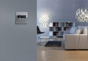 GIRA präsentiert ein Wohnzimmer mit einer hellgrauen Schaltuhr mit Display an einer hellgrauen Wand.