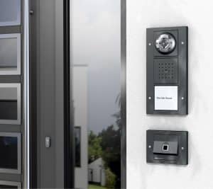 GIRA präsentiert eine dunkelgraue Türstation mit Kameramodul und Gegensprechanlage, darunter ein Fingerprint-Türöffner neben einer modernen Eingangstüre.