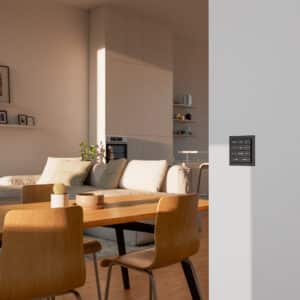 Ein Gira 4fach-Schalter F200 flach TS4 70 4F in Anthrazit bzw. Schwarz mit Steuerungstasten für Heizung, Jalousie, Downlights und für Wohn- und Eingangsbereich, montiert an einer hellen Wand im modernen Wohnbereich.