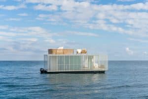 Seitenansicht des schwimmenden Pavillons auf offener See mit Sicht auf den Motor, der Außenverkleidung aus senkrechten weißen Sprossen und der Reling am Oberdeck von Gira.