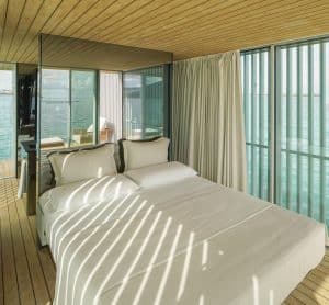 Schlafbereich mit Doppelbett in weißem Bezug und verspiegelter Rückwand, heller holzverkleiderter Decke, passendem Dielenboden und bodentiefer Verglasung an der Seite mit beigem Vorhang von Gira.