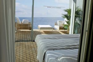 Sicht vom Schlafbereich auf die Deckterrasse mit Outdoormöbeln, welche einer Aussichtsplattform aufs Meer gleicht, betretbar durch großzügige Glasschiebetüren bereitgestellt von Gira.
