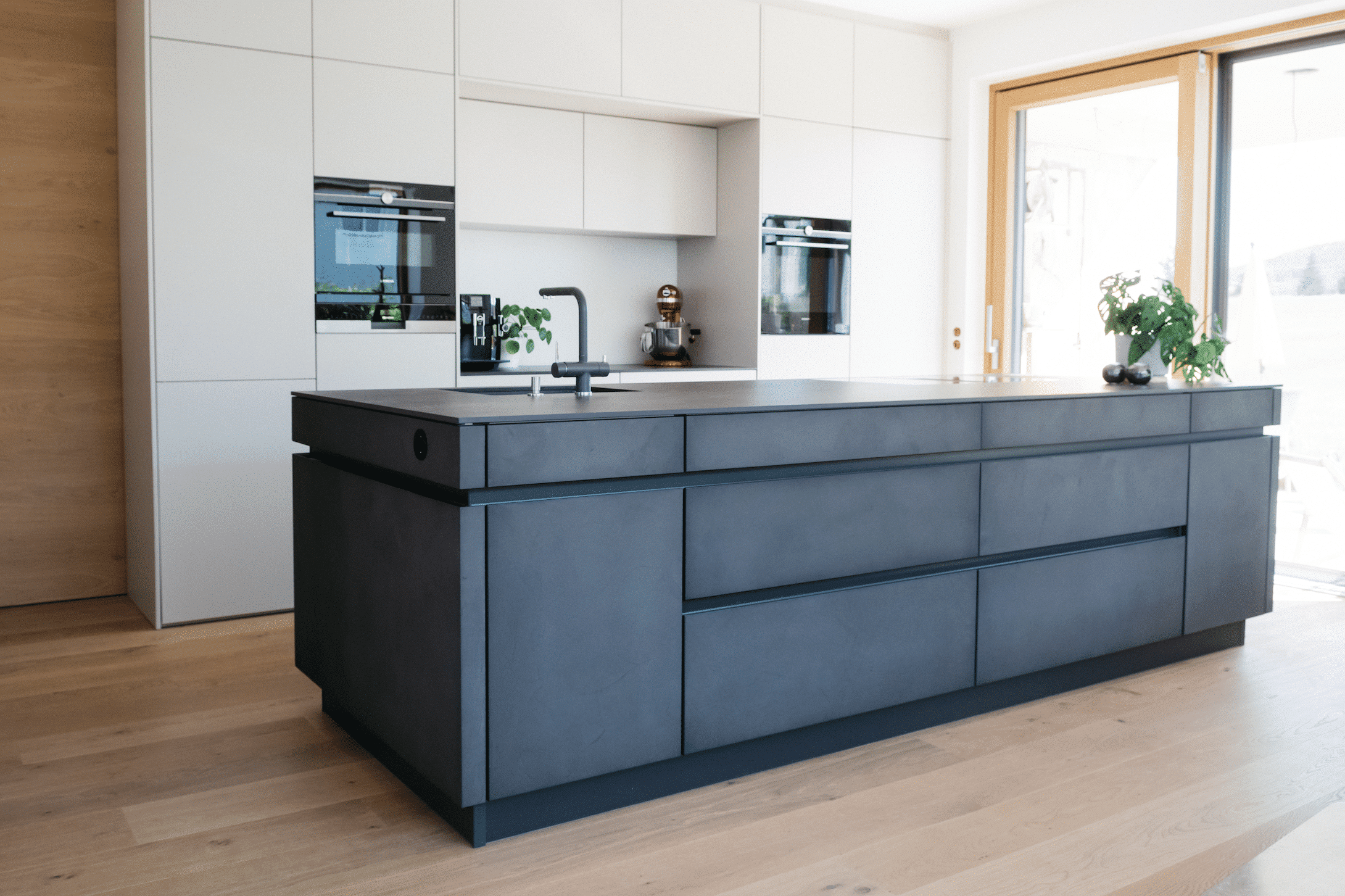 Moderne Küche mit Kochinsel aus dunkler Betonoptik und Wandverbau mit weißen Fronten von WOHNHAUS Grill & Ronacher.