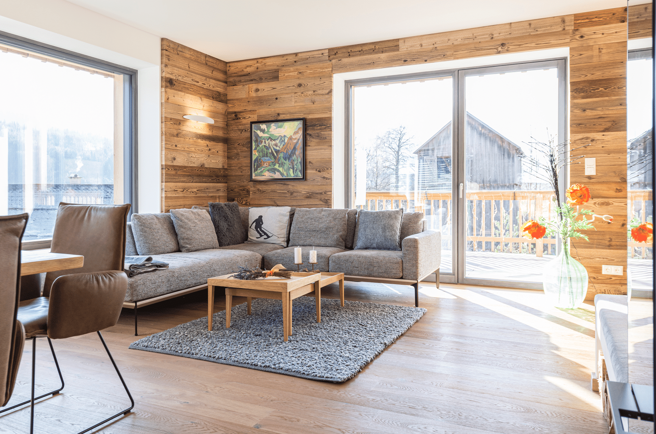 Gemütliches Wohnzimmer mit rustikaler Holzrückwand, grauer Couch und Holzbeistelltisch auf grauem Teppich und Holzboden von WOHNHAUS Grill & Ronacher.