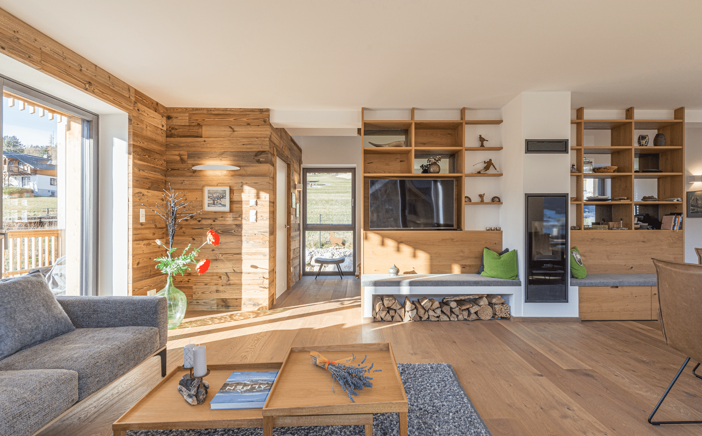 Gemütliches Wohnzimmer mit Rückwänden aus Vollholz, Sitzbank neben integriertem Holzofen, grauer Couch und Stauraum in den Holzregalen von WOHNHAUS Grill & Ronacher.