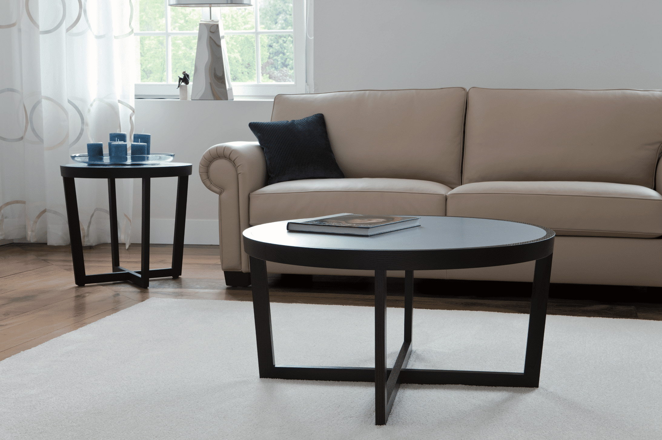 Grill & Ronacher zeigt eine beige Ledercouch mit kleinem blauem Polster, einem runden Holztisch und Beistelltisch mit Kerzen darauf von Bielefelder Werkstätten.