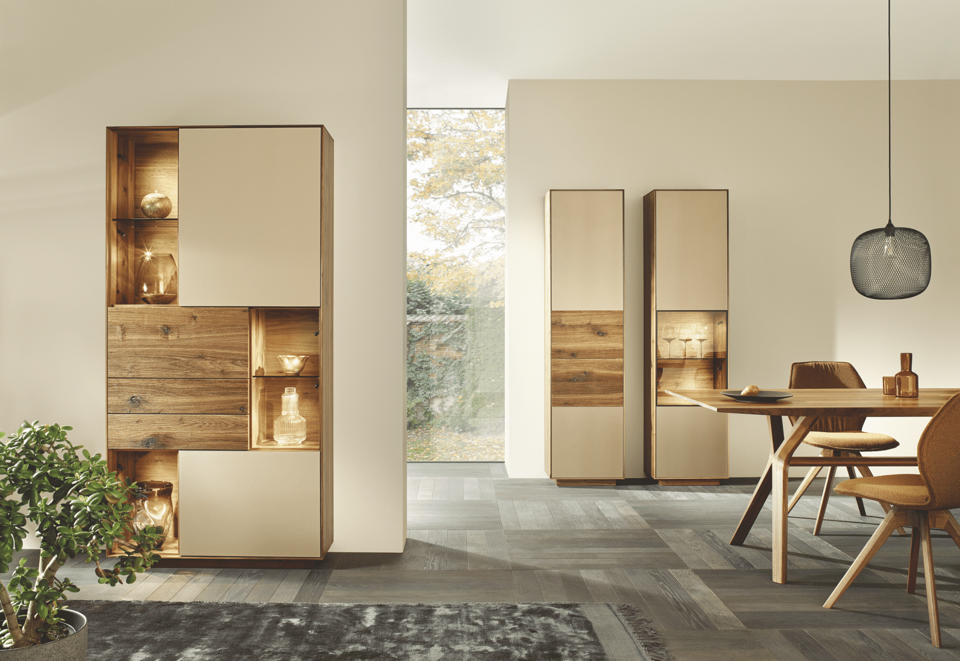 WOHNHAUS Grill & Ronacher zeigt ein Esszimmer mit einem Holztisch und einer runden Hängelampe darüber und einer beleuchteten Vitrine an der Wand und zwei hohen Regalen von Anrei.