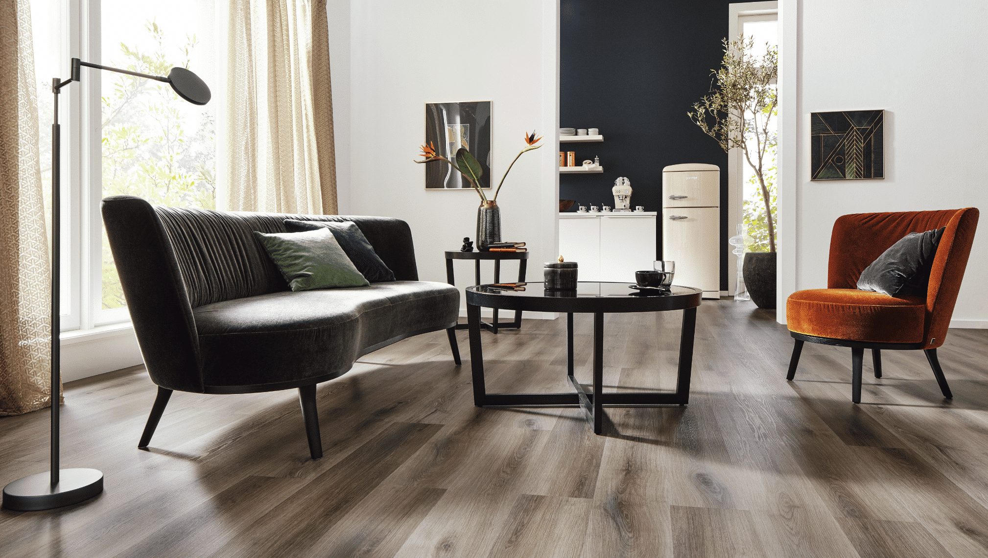 WOHNHAUS Grill & Ronacher zeigt ein Wohnzimmer mit einer braunen Couch, einem orangen Polsterstuhl und rundem Beistelltisch aus Glas und Holz in der Mitte von Bielefelder Werkstätten.