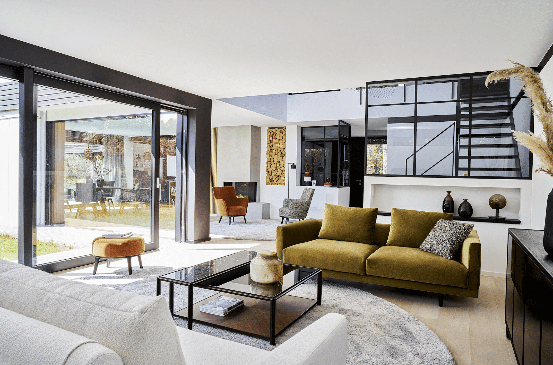Blick in den offenen Wohnraum einer Villa mit einem Samtsofa in gold, Polsterstühlen in orange und einem schwarzen Couchtisch von Bielefelder Werkstätten präsentiert von Grill & Ronacher.