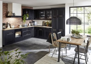 WOHNHAUS Grill & Ronacher zeigt eine L-förmige Küche im Landhausstil mit schwarzen Fronten, beiger Arbeitsfläche und goldenen Griffleisten mit angeschlossenen Essbereich von Nobilia vor einer Ziegelwand.