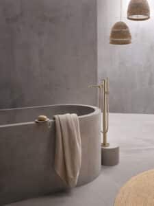 Hansa zeigt eine moderne freistehende Badewanne mit einer freistehenden Armatur in gold mit obenliegendem Hebel und mit stabförmigen Handdusche, auf einem Sockel montiert.