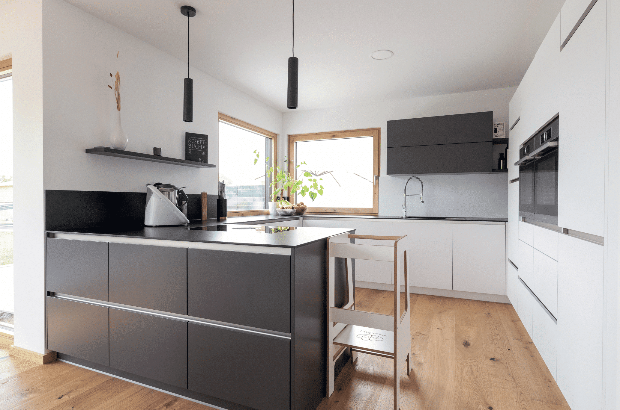 Hartl Haus zeigt eine Küche mit Holzboden und einer schwarzen und weissen Küchenzeile in U-Form, einem großen Wandschrank mit Geräten und schwarzen Hängeschränken über dem Spülbecken mit silberner Armatur.