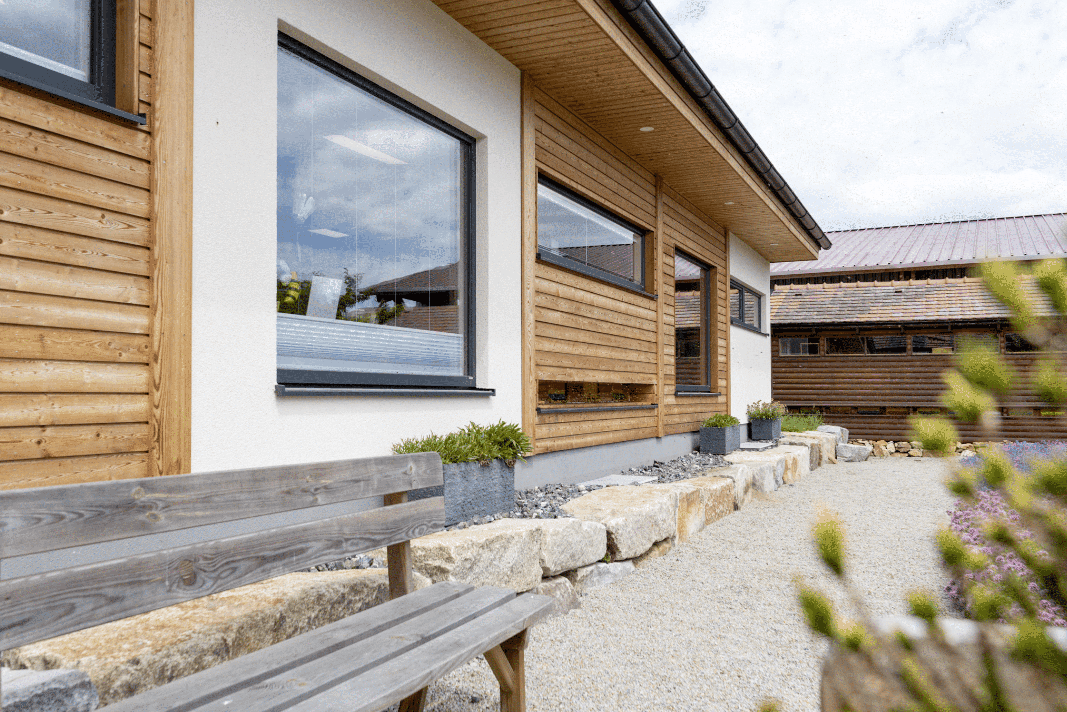 HARTL HAUS zeigt einen Bungalow in weiß mit Holzverbau, ein quadratisches Fenster, eine Steinmauer und Bänkchen.