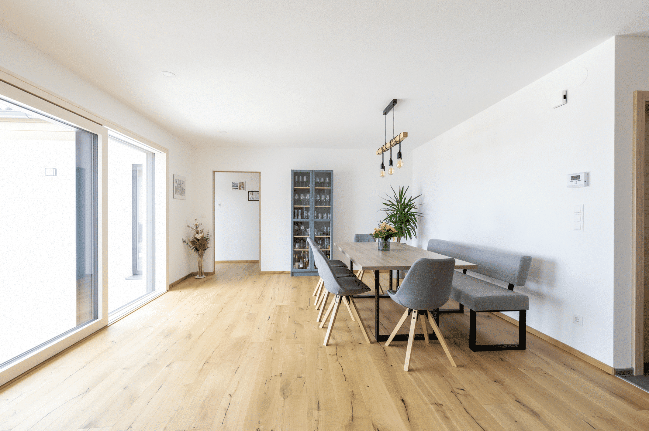 HARTL HAUS zeigt ein Esszimmer mit hellem Holzboden, grauen Polstermöbeln, einem Holztisch und blauer Glasvitrine.