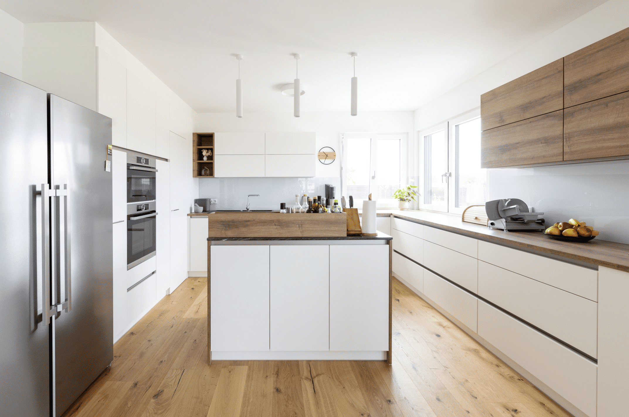 HARTL HAUS zeigt eine weiß matte, grifflose Küche mit Holzelementen, großem Kühlschrank und Insel in der Mitte.