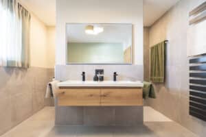 HARTL HAUS zeigt ein Badezimmer mit großformatigen Boden- und Wandfliesen in Betonoptik, einem schwebenden Waschtisch mit Holzschubladen, jeweils einer Dusch- und einer WC-Nische und einem schwarzen Handtuchwärmer in Sprossenoptik.