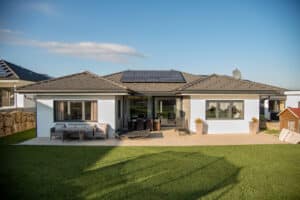 HARTL HAUS zeigt einen Bungalow mit Walm-Kehldach-Kombination, dachmontierten Solarpaneelen, Gartenfläche und einer gefliesten Terrasse mit Outdoormobiliar.