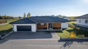 HARTL HAUS zeigt einen Bungalow mit Kombinationsdach, Gartenfläche, Doppelgarage und einem überdachten Eingangsbereich mit Holzfassade und Sitzgelegenheiten.
