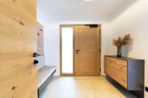 HARTL HAUS zeigt den Vorraum eines Hauses mit Garderobe bestehend aus einer schwebenden Bank, Wandpaneel zur Aufhängung von Kleidungsstücken und einer Hauseingangstüre aus Holz mit Seitenlicht und schwarzer Klinke im gleichen Design wie die Innentüre.