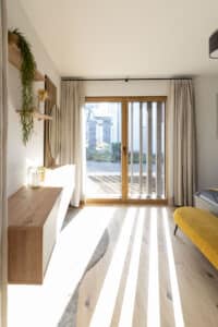 Schlafzimmer mit Naturparkett, Holzterrassentür, wandmontiertem Schwebeboard mit Holzsprossenpaneel, Spiegel und schwarzer Vorhangstange mit beigem Vorhang von HARTL HAUS.