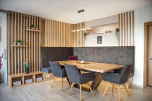 HARTL HAUS zeigt eine Essecke mit gepolsterter Sitzbank, Esstisch, 3 Polsterstühlen und indirekt beleuchteten Holzsprossen-Wandpaneelen.