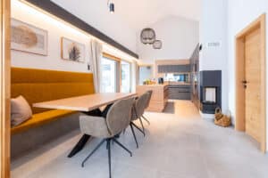 Offene Wohnküche mit schwebender gepolsterter Sitzbank, Esstisch, 3 Polsterstühlen, dahinter eine c-förmige Inselküche, ein gemauerter Kamin und eine Terrassenschiebetür von HARTL HAUS.